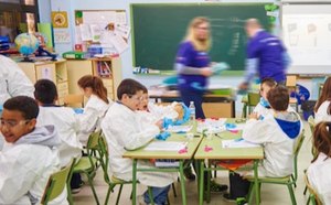 A handful of students sit at their desks wearing lab coats and gloves. Two staff volunteers walk around to support students. 