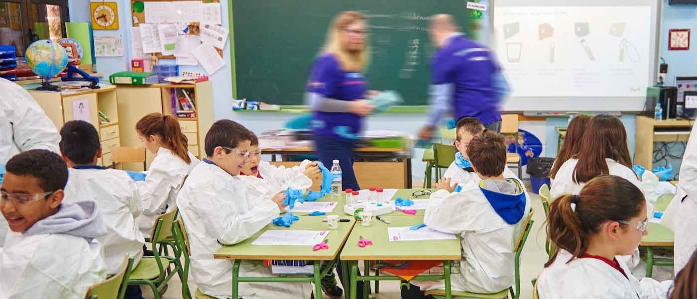 A handful of students sit at their desks wearing lab coats and gloves. Two staff volunteers walk around to support students.