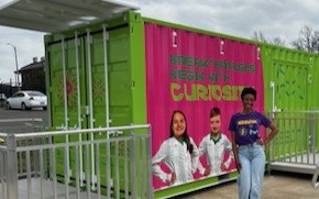 Danielle Chavis wears a SPARK t-shirt and stands in front of the Curiosity Cube which is bright green, has images of two young scientists with the words “Breakthroughs Begin with Curiosity” on the front. 