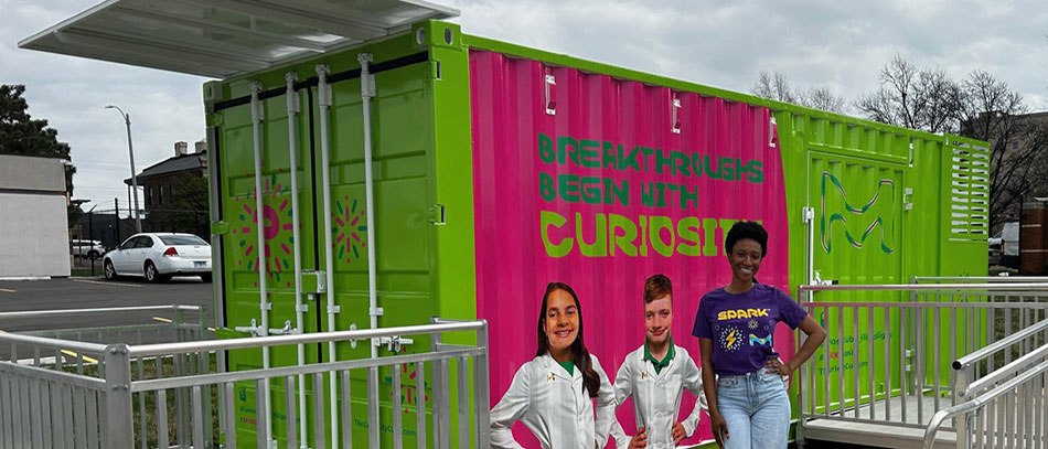 Danielle Chavis wears a SPARK t-shirt and stands in front of the Curiosity Cube which is bright green, has images of two young scientists with the words “Breakthroughs Begin with Curiosity” on the front.