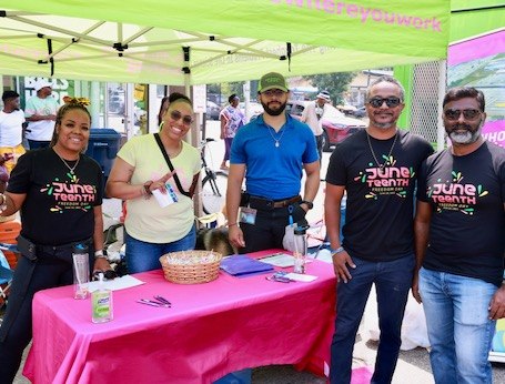 A group of employees stand together at a community event. They are standing under a company tent and smiling at the camera. 