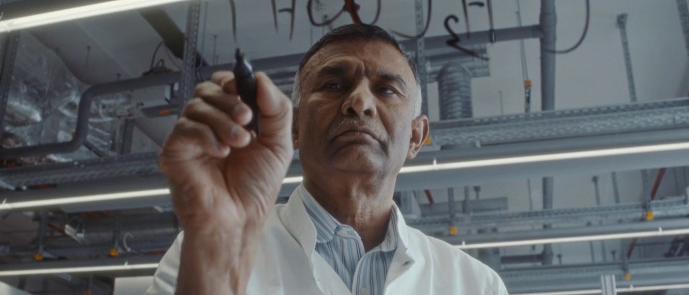 Dr. Ponnusamy wears a lab coat is writes out chemical compounds on glass with a marker.