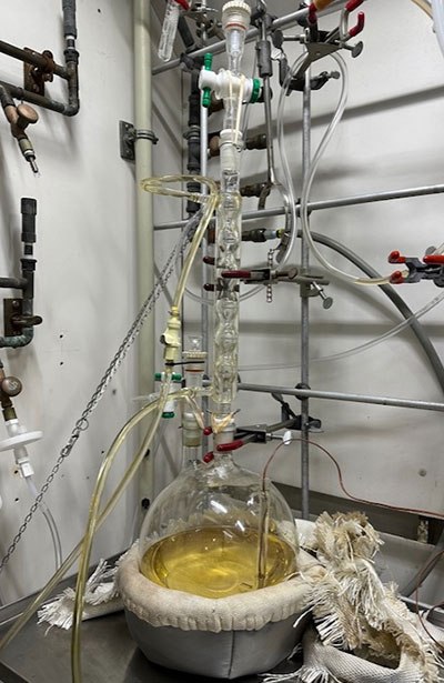 Round-bottom flask sits on a lab bench with tubes coming out in every direction. The liquid inside appears to be an off-yellow color.