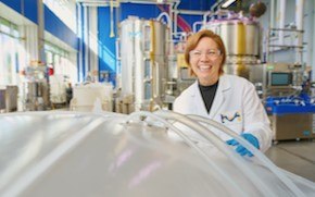 Shannon wears a lab coat, blue gloves and safety glasses and stands in an M Lab — a non-GMP biomanufacturing setting. She is smiling and holding onto a bin bag, which is situated in the lower left corner.