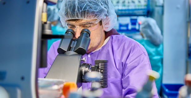 Scientist wears a purple lab coat, protective eyewear and a hair net while peering into a microscope. 