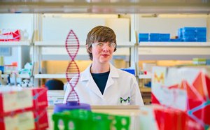 实验室场景中，一名身着白大褂的研究人员伫立于摆满各类实验器材的货架前。前景处陈列着DNA双螺旋结构模型，色彩斑斓的盒子清晰可见，暗示着不同产品或样本的存在。