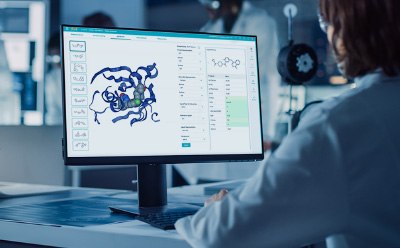 A scientist in a lab coat is seated at a desk, interacting with a computer screen displaying a molecular structure. The screen shows a 3D representation of a protein or complex, with various tools and data options visible on the side.