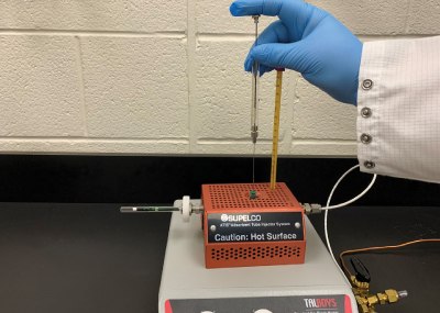 Right Hand wearing a blue glove shown adding terpene test mix using Supelco® Adsorbent Tube Injector System (ATIS) to the Carbotrap® T420 tube kept on a platform against a white colored wall.