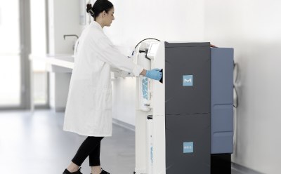 : A woman in lab coat and blue protective gloves moves the Milli-Q CLX 8Series clinical water system to a space against a wall.